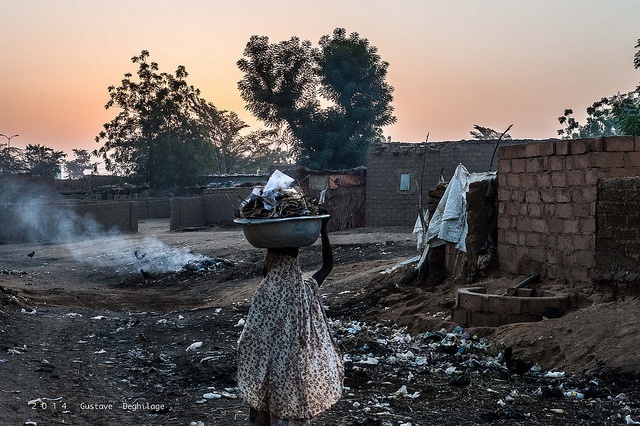 At sunset a woman in a village in Niger carries a basket of food on her head, in the background are brick houses and trees